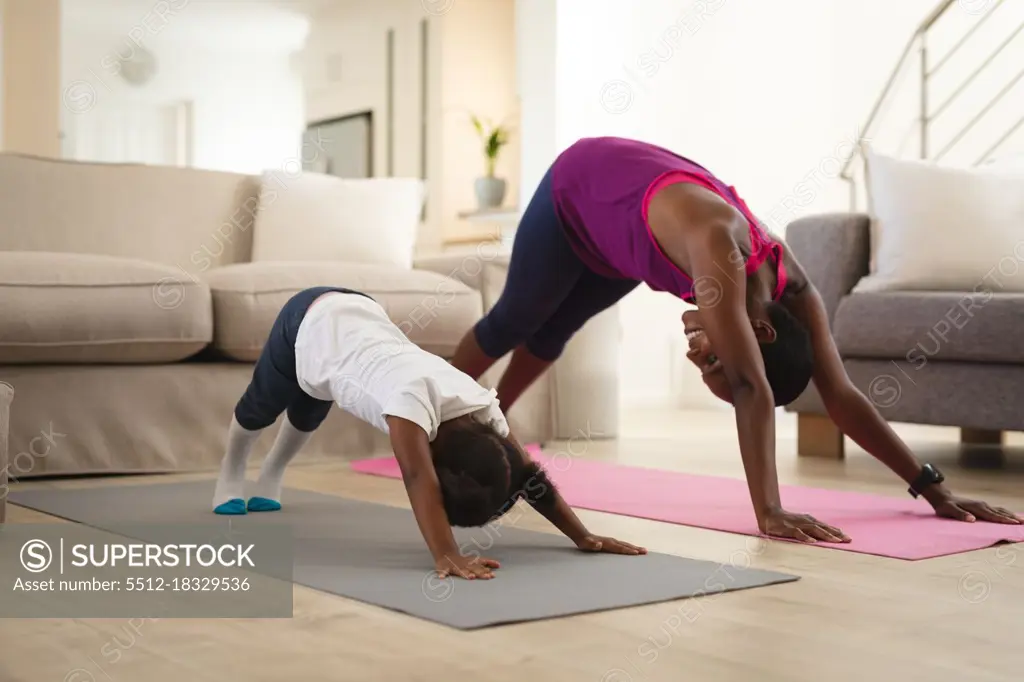 Smiling african american mother and daughter practicing yoga, stretching on mats in living room. family spending time together at home.