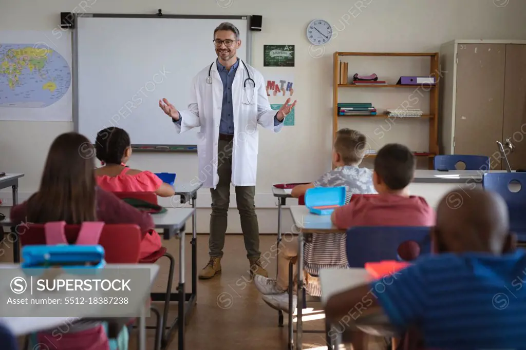 Caucasian male doctor talking to group of diverse students sitting in the class at school. health protection and safety at school during covid-19 pandemic concept