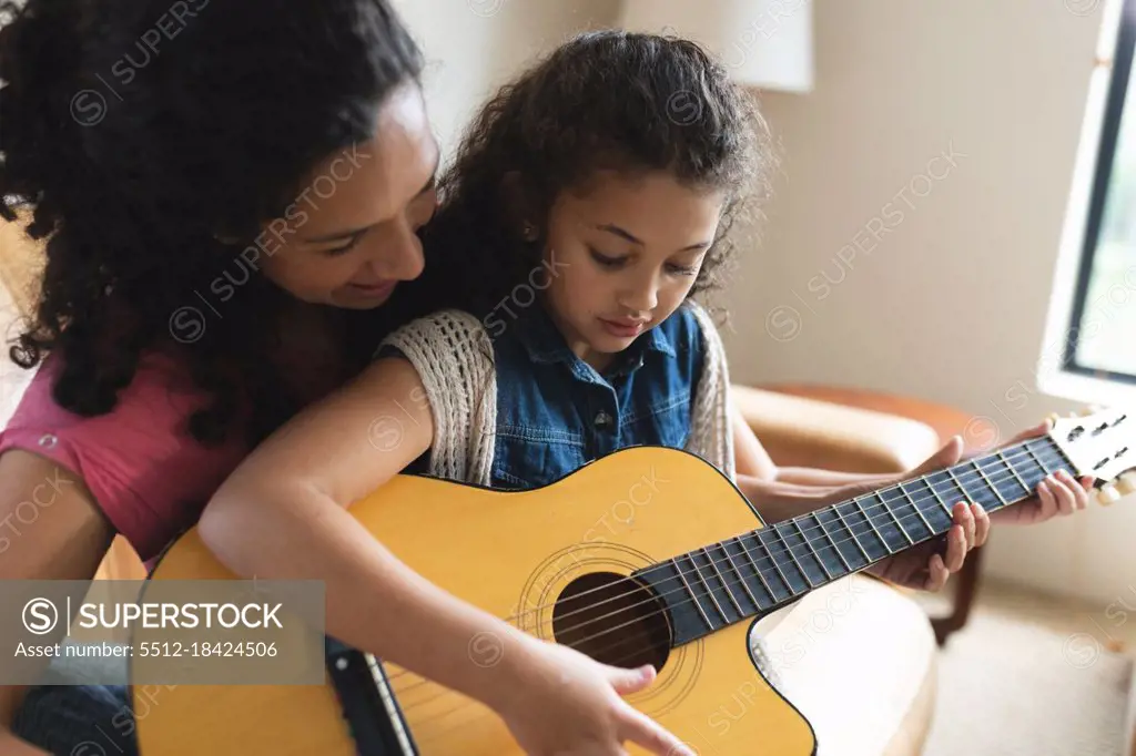 Mixed race mother and daughter sitting on sofa and playing guitar. domestic lifestyle and spending quality time at home.