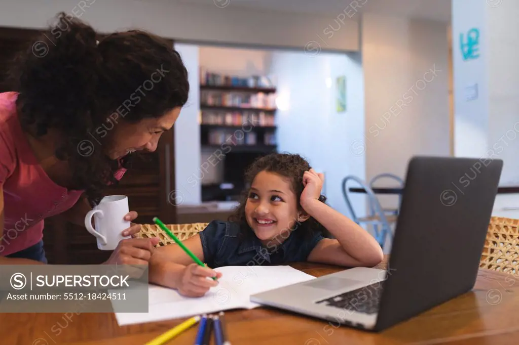 Smiling mixed race mother holding coffee, helping her daughter do homework. domestic lifestyle and spending quality time at home.