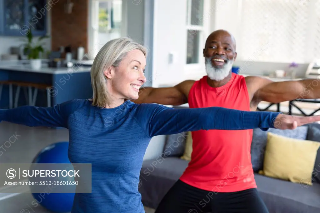 Happy senior diverse couple in exercise clothes practicing yoga together, stretching. healthy, active retirement lifestyle at home.