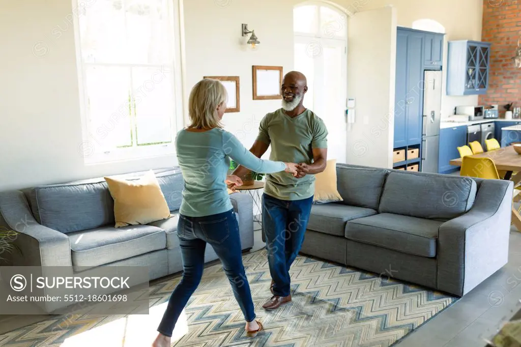 Happy senior diverse couple in living room dancing together. retirement lifestyle, spending time at home.