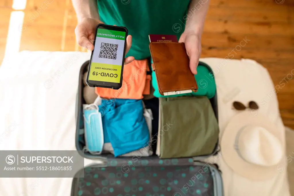 African american albino man packing and holding smartphone with covid 19 vaccine passport. holiday and travel preparation during covid 19 pandemic.