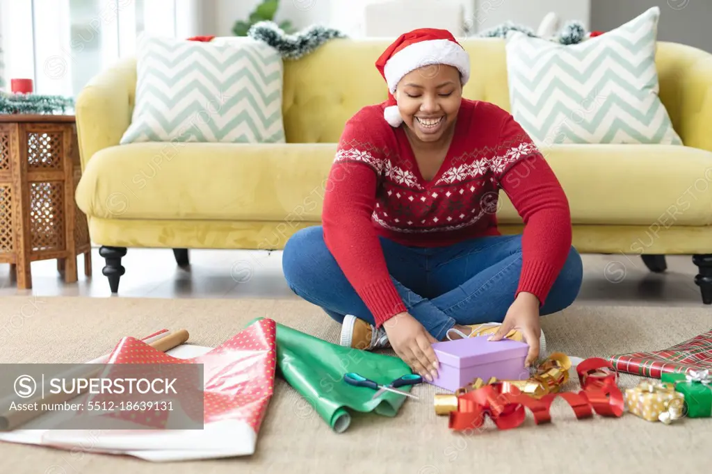 Happy african american plus size woman in santa hat wrapping presents at home. christmas, festivity and tradition concept.
