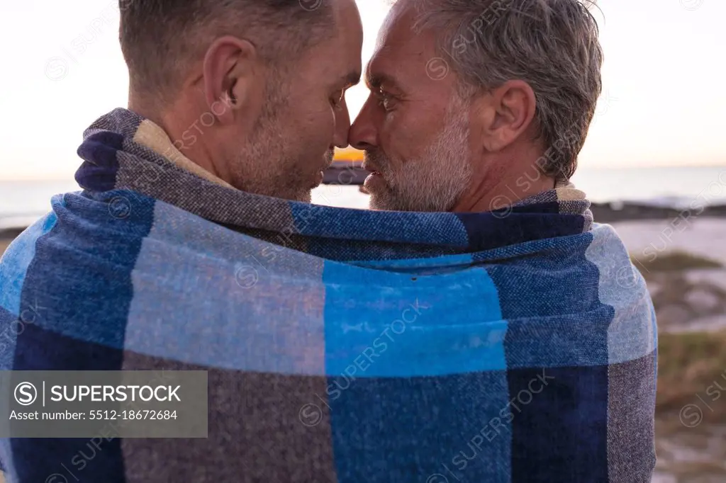 Happy caucasian gay male couple wrapped in blanket, embracing and taking selfie on beach at sunset. summer road trip and holiday in nature.