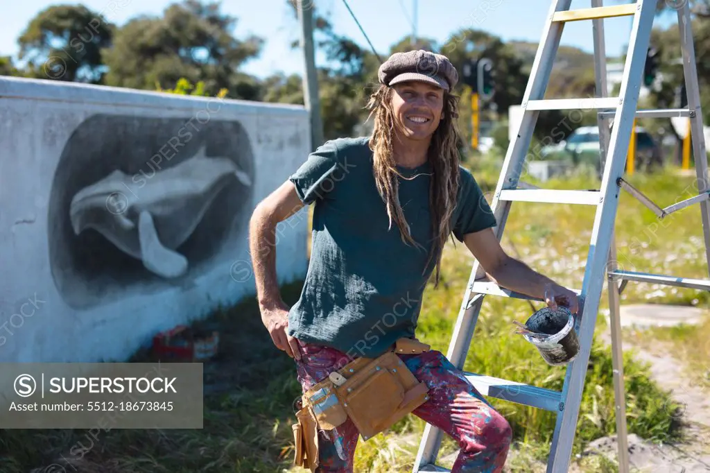 Portrait of smiling male artist leaning on ladder in front of mural painting of whale on wall. street art and skill.
