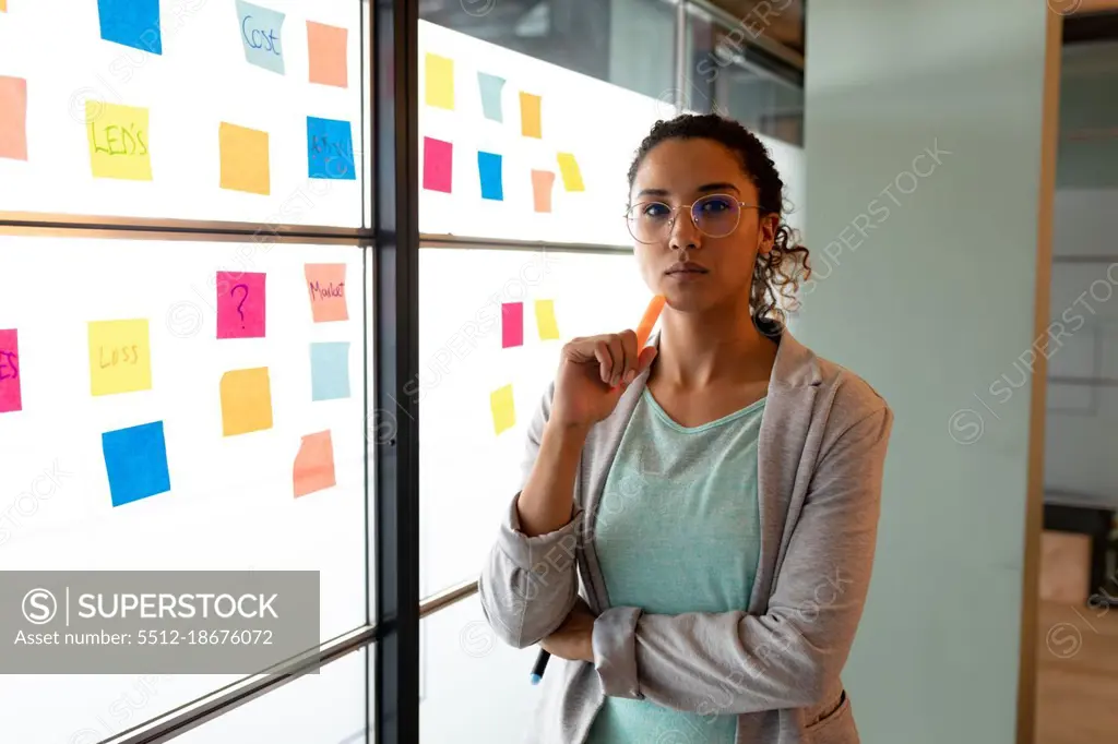 Portrait of confident biracial businesswoman in smart casuals by sticky notes on wall in office. creative business, modern office and business plan.