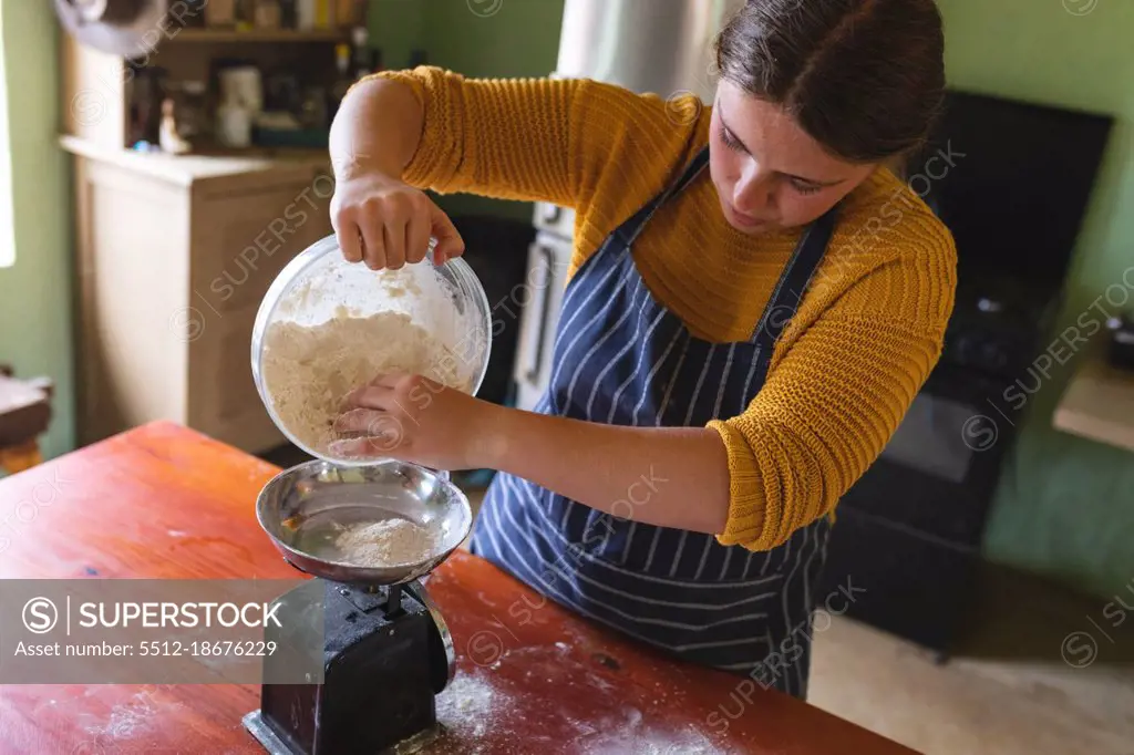 Young woman pouring flour from container on weight scale at table in kitchen. domestic lifestyle and healthy eating.