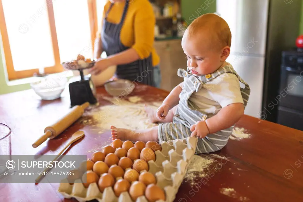 Cute baby playing with egg carton on wooden table while mother preparing food in kitchen. innocence, family and healthy eating.
