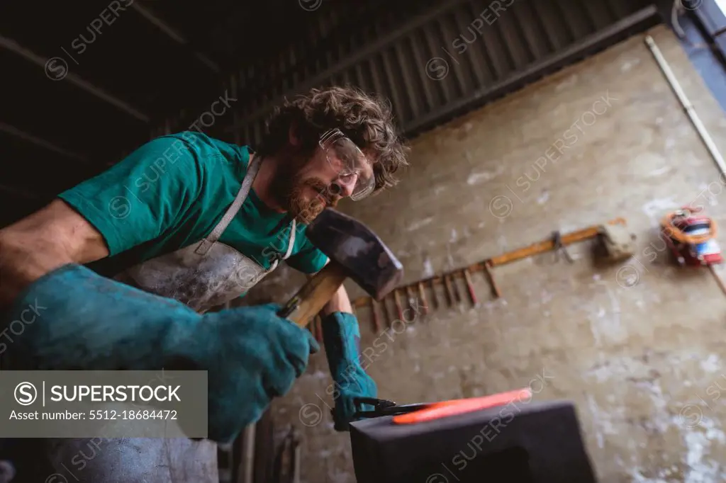 Bearded caucasian blacksmith in protective workwear forging with hammer on anvil in industry. forging, metalwork and manufacturing industry.