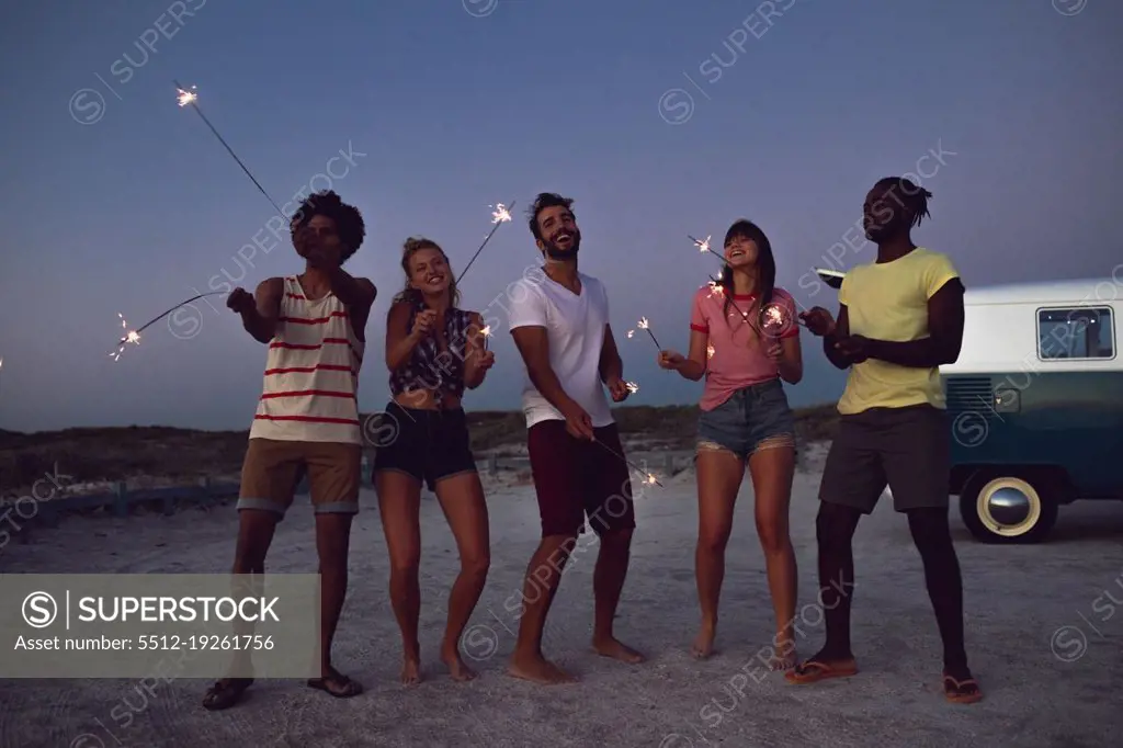 Front view of happy group of diverse friends playing with sparklers on the beach at dusk