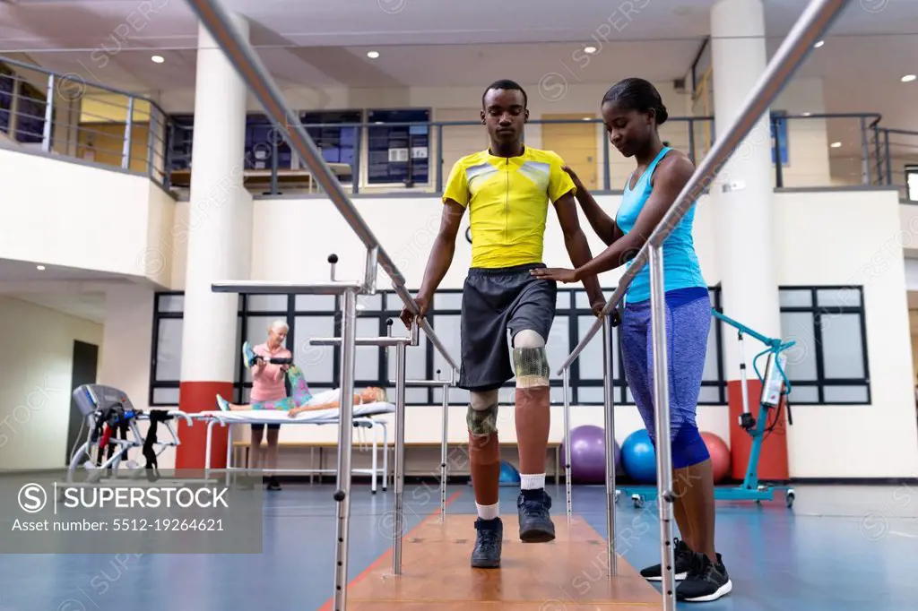 Low angle view of African-american physiotherapist helping disabled African-american man walk with parallel bars in sports center. Sports Rehab Centre with physiotherapists and patients working together towards healing