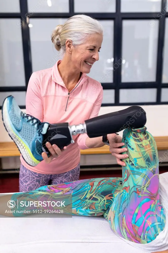 Front view of Happy Caucasian active senior woman assisting disabled Caucasian woman to stretching leg in sports center. Sports Rehab Centre with physiotherapists and patients working together towards healing