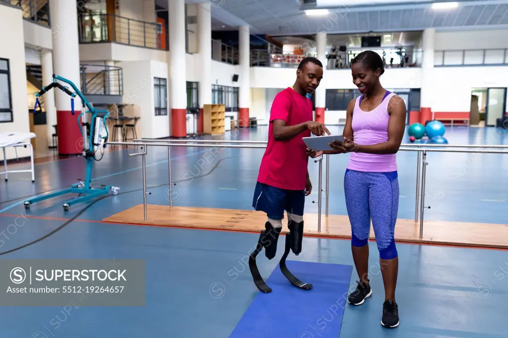 Front view of African-american female trainer and disabled African-american man discussing over digital tablet in sports center. Sports Rehab Centre with physiotherapists and patients working together towards healing