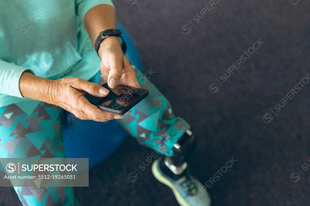 Close-up of disabled active senior Caucasian woman with leg amputee using mobile phone while sitting on exercise ball in fitness center. Strong active senior female amputee training and working out