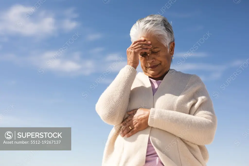 Low angle view of tensed biracial senior woman with hand on forehead at beach against blue sky. lifestyle and negative emotion.