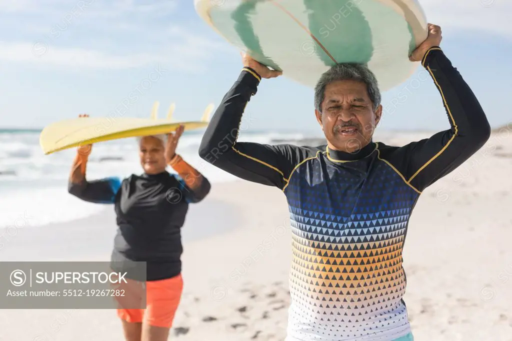 Multiracial senior couple enjoying retirement together while carrying surfboards over heads at beach. water sport and active lifestyle.