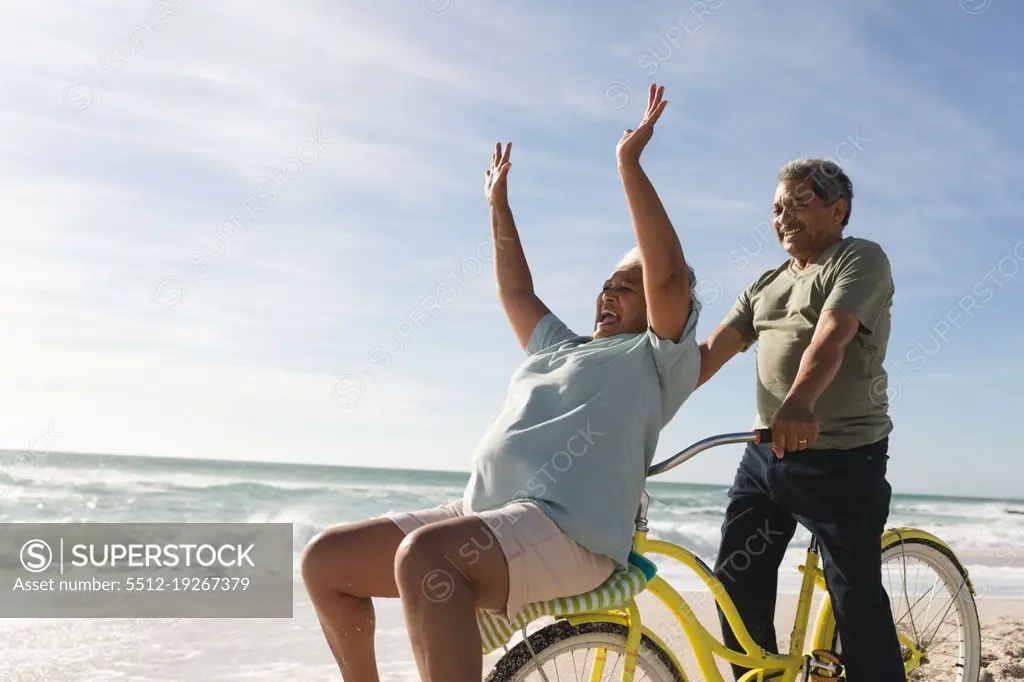 Cheerful biracial senior woman cheering while man riding bicycle at beach on sunny day. active lifestyle and transportation.
