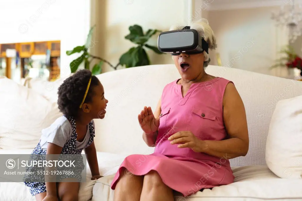 African american girl looking at her grandmother wearing vr headset sitting on the couch at home. virtual reality and futuristic technology concept, unaltered.
