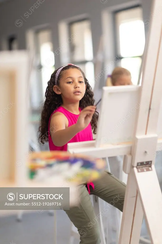 Cute biracial elementary schoolgirl painting on easel during drawing class in school. unaltered, childhood, education, art, painting, activity and back to school concept.