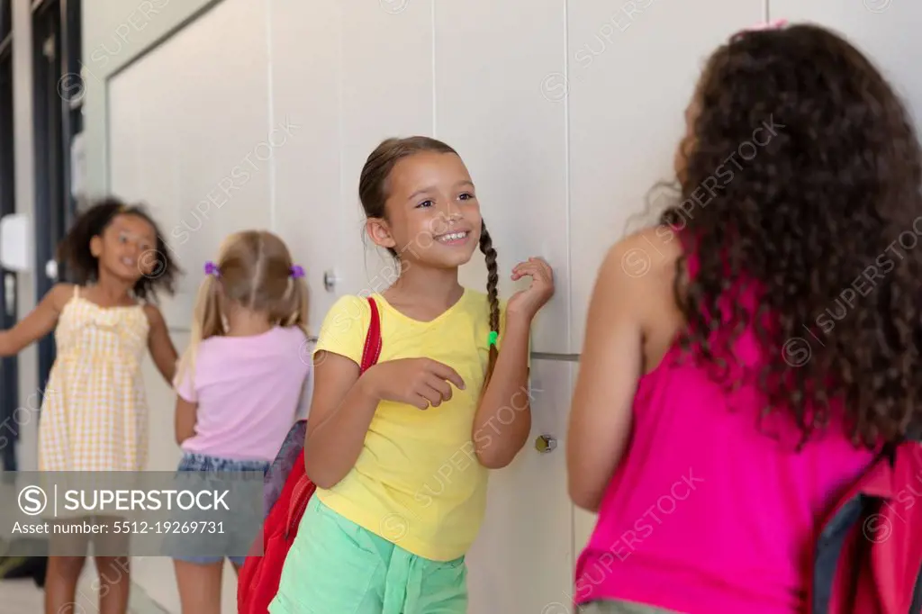 Multiracial elementary schoolgirls talking while standing by lockers in school corridor. unaltered, childhood, together, communication, education and back to school concept.