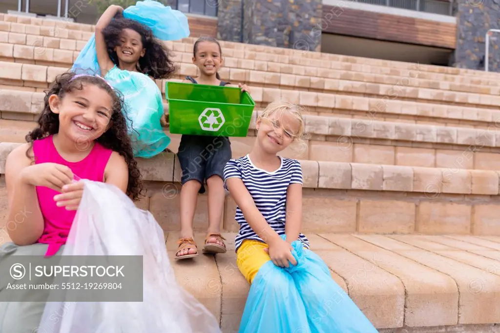 Portrait cheerful multiracial elementary schoolgirls with garbage bags sitting on school steps. unaltered, sustainable lifestyle, education, cleaning, responsibility and recycling concept.