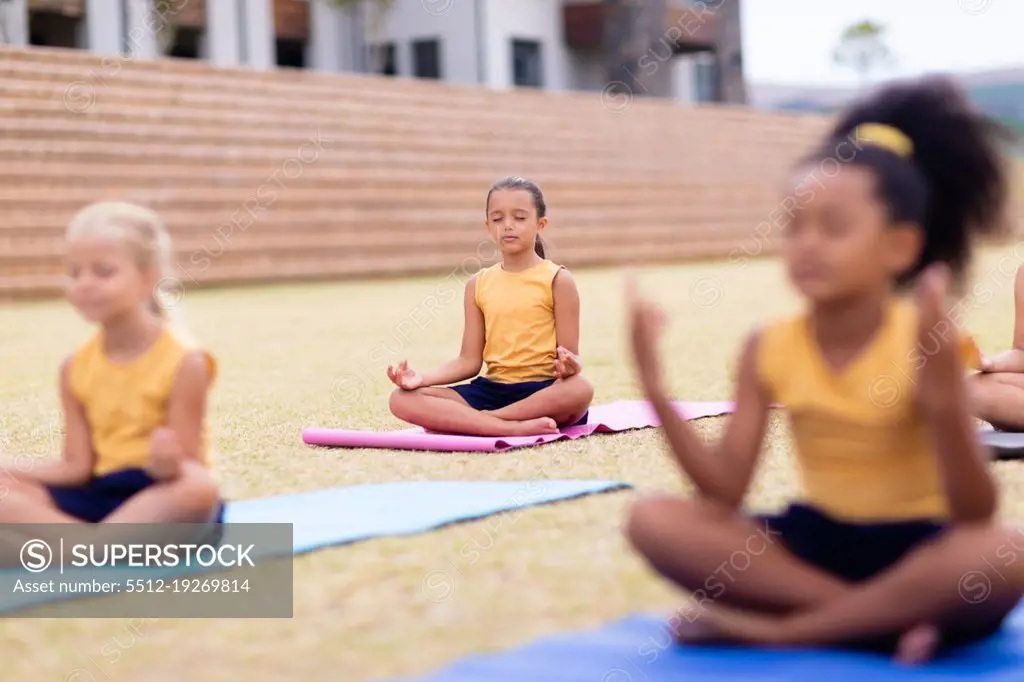 Multiracial elementary schoolgirls meditating while sitting on yoga mat at school ground. unaltered, childhood, education, activity, sports training, yoga and physical education concept.