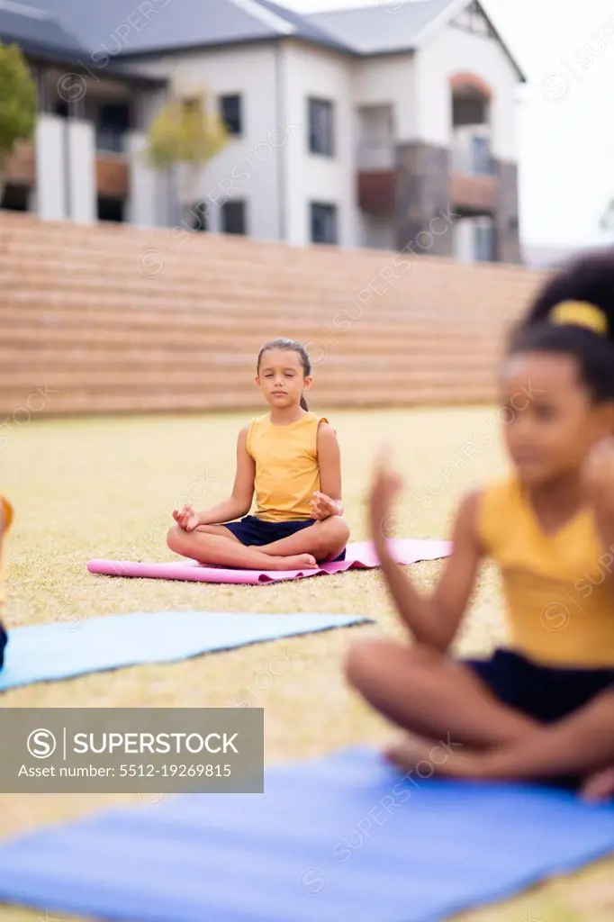 Multiracial elementary schoolgirls meditating while sitting on yoga mat against school. unaltered, childhood, education, activity, sports training, yoga and physical education concept.