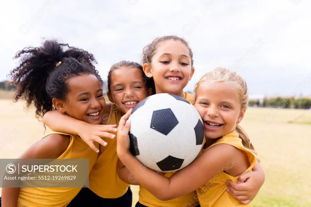 Happy multiracial elementary schoolgirls with soccer ball embracing while standing on ground. unaltered, childhood, education, together, sports training and sports activity concept.