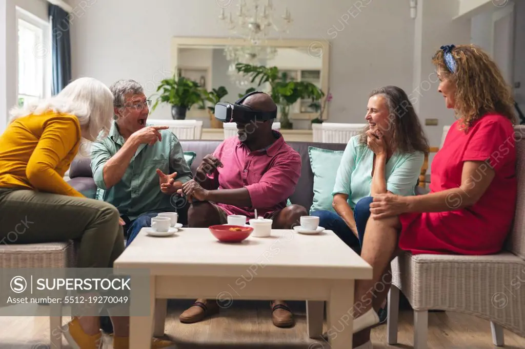 Happy multiracial senior man and women looking at male friend using vr headset at home. unaltered, lifestyle, social gathering, friendship, virtual reality, futuristic and modern technology.