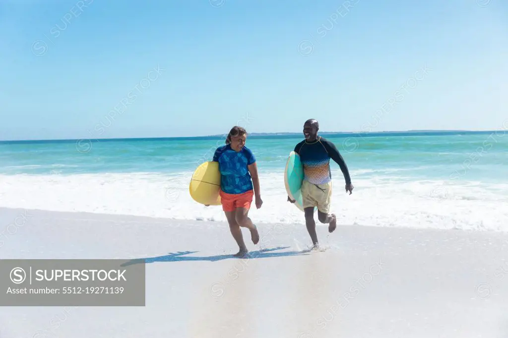African american senior couple carrying surfboards running at beach against blue sky with copy space. unaltered, togetherness, active lifestyle, aquatic sport and holiday concept.