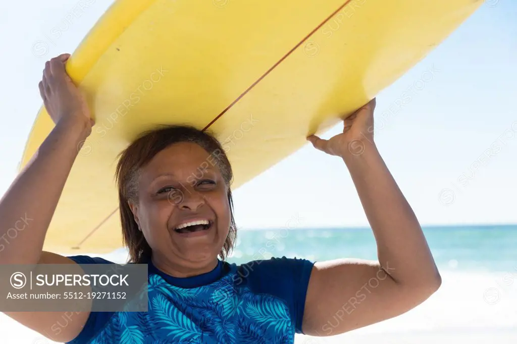 Portrait of happy retired african american senior woman carrying yellow surfboard on head at beach. unaltered, active lifestyle, aquatic sport and holiday concept.