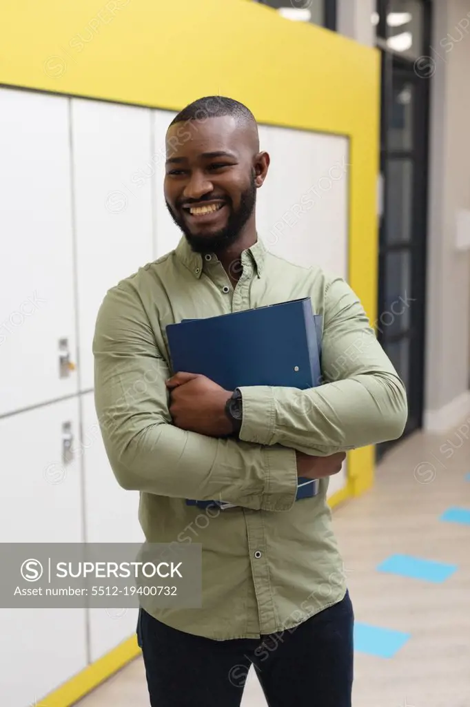 Smiling african american young male teacher with files looking away while standing in corridor. unaltered, education, occupation and school concept.