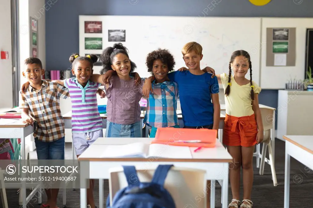 Portrait of smiling multiracial elementary school students standing with arm around in classroom. unaltered, education, childhood, learning, together and school concept.