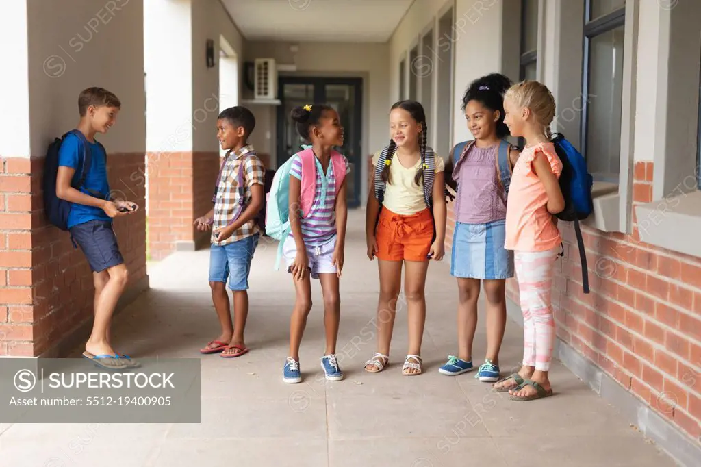 Full length of multiracial elementary school students talking while standing in school corridor. unaltered, education, childhood, together and school concept.
