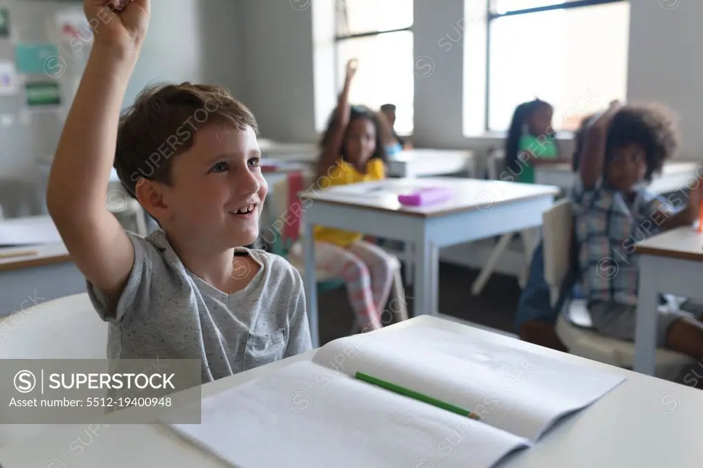 Caucasian elementary schoolboy raising hand while sitting with multiracial classmates in background. unaltered, education, childhood, learning, studying, intelligence, book and school concept.