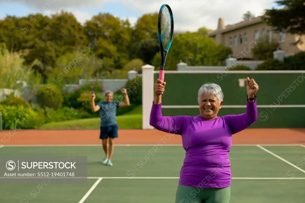 Happy biracial senior woman holding racket and gesturing at tennis court with man in background. achievement, winning, unaltered, sport, competition, retirement, healthy and active lifestyle.