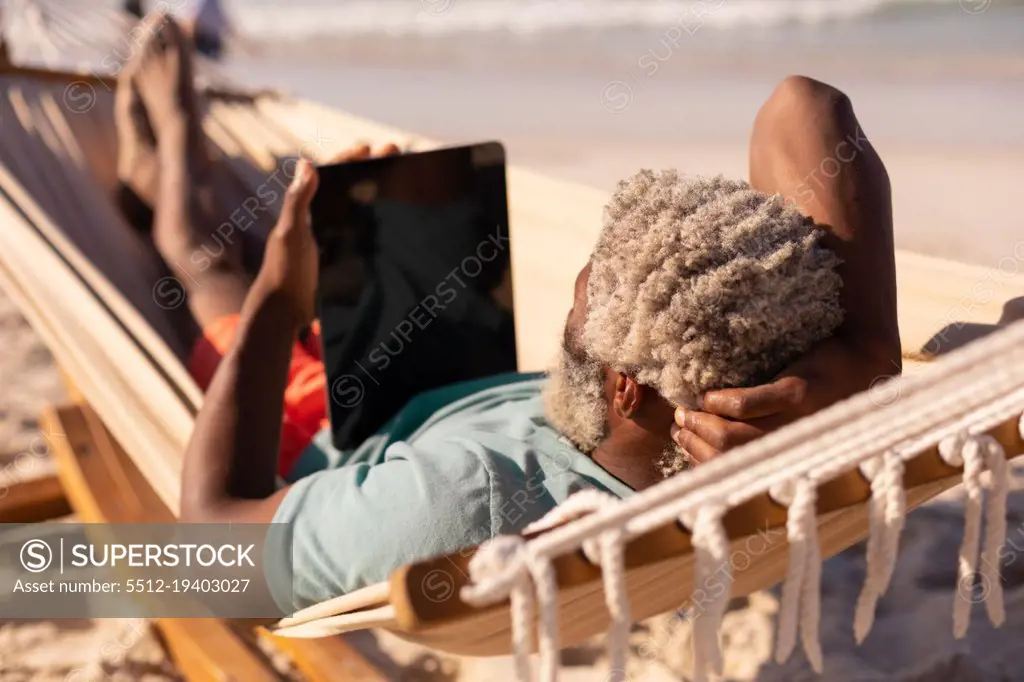 African american senior man with gray hair using digital tablet while lying on hammock at beach. summer, copy space, technology, relaxation, unaltered, lifestyle, retirement, enjoyment, holiday.