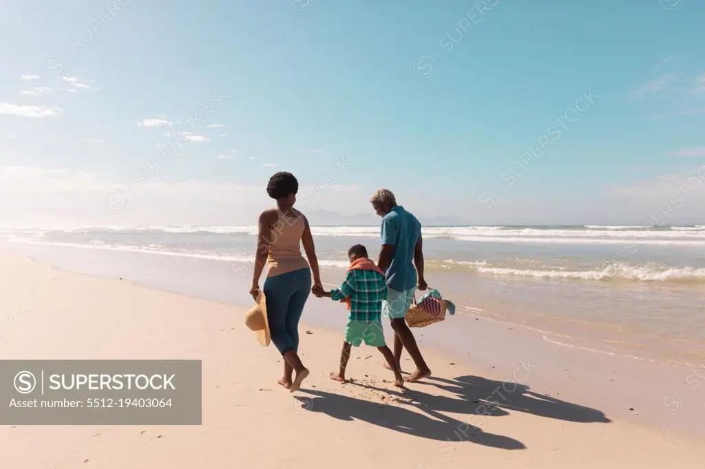 African american grandparents holding boy's hands and walking at sandy beach against sky in summer. nature, copy space, unaltered, family, love, togetherness, childhood, enjoyment, holiday concept.