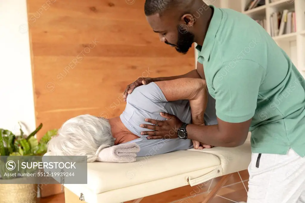 African american male physiotherapist giving back massage therapy to caucasian senior woman at home. Physiotherapy and rehabilitation concept