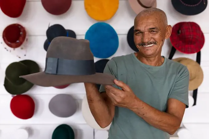 Portrait close up of a senior mixed race man smiling and holding a finished hat in the showroom at a hat factory, with various hats on display in the background