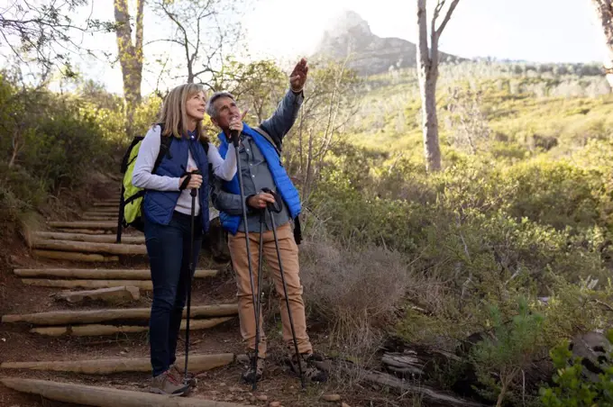 Front view of a mature Caucasian woman and man wearing backpacks and using Nordic walking sticks, stopping on a downhill trail to admire the view and pointing to the distance during a hike in the countryside