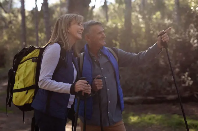 Side view close up of a mature Caucasian woman and man wearing backpacks and using Nordic walking sticks, stopping on a trail to admire the view and pointing to the distance during a hike in the countryside