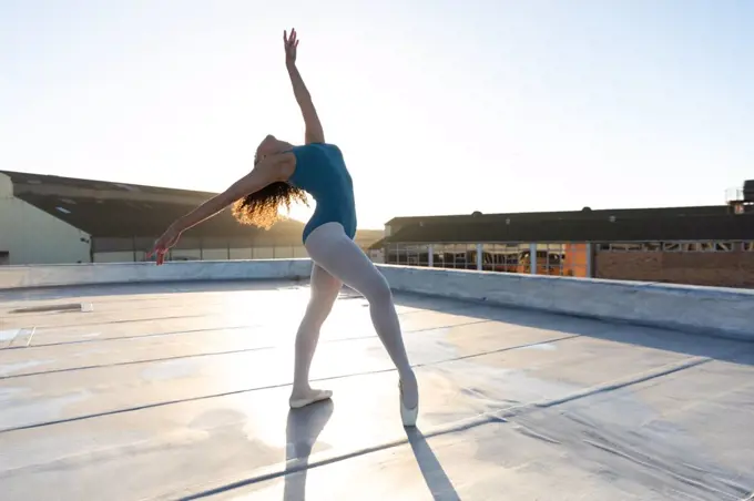 Side view of a young mixed race female ballet dancer leaning back in a ballet pose with arms oustretched, on the rooftop of an urban building, backlit by sunlight