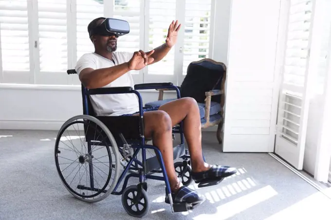 A senior retired African American man at home, sitting in a wheelchair in his underwear in front of a window on a sunny day using a VR headset with his arms outstretched in front of him, self isolating during coronavirus covid19 pandemic