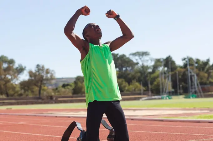 Fit, mixed race disabled male athlete at an outdoor sports stadium, kneeling on race track after race with arms in the air wearing running blades. Disability athletics sport training.