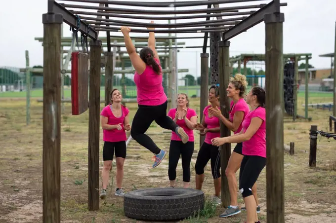 Multi-ethnic group of women all wearing pink t shirts at a boot camp training session, exercising, hanging from monkey bars. Outdoor group exercise, fun healthy challenge.