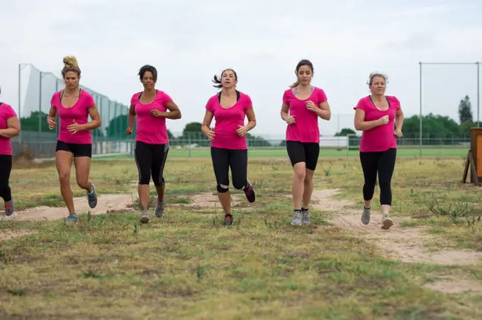 Multi-ethnic group of women all wearing pink t shirts at a boot camp training session, exercising, running on a field. Outdoor group exercise, fun healthy challenge.
