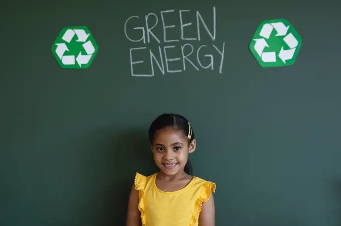 Front view of smiling schoolgirl standing against green energy board in classroom of elementary school