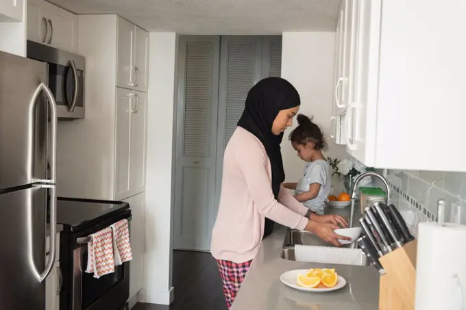 Side view of mixed race mother wearing hijab working in kitchen room while daughter sitting behind her at home